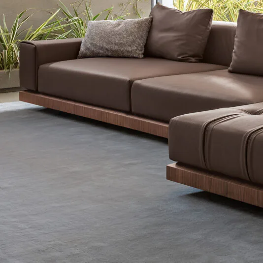 A sophisticated living room corner showcases the Contemporary High End Modular Corner Sofa featuring a luxurious brown leather finish and adorned with a textured gray throw pillow. The furniture is arranged against a backdrop of potted plants and a concrete wall, with natural light emphasizing its sleek lines and minimalist design.