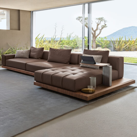 A modern living room showcases a Contemporary High-End Modular Corner Sofa, equipped with plush cushions and an integrated wooden side table supporting a laptop. The space is illuminated by floor-to-ceiling windows that reveal views of an outdoor garden and distant mountains, all complemented by minimalist decor.