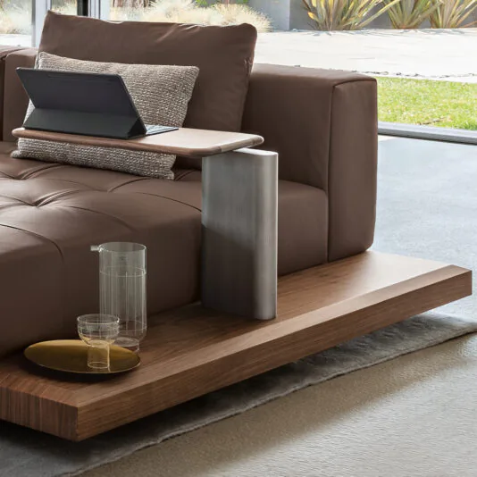A modern living room features the Contemporary High End Modular Corner Sofa with a built-in wooden side table. On the table, there is a tablet on a stand, a textured pillow, a glass jug, and two small glasses on a tray. The background showcases large windows with views of an outdoor garden.