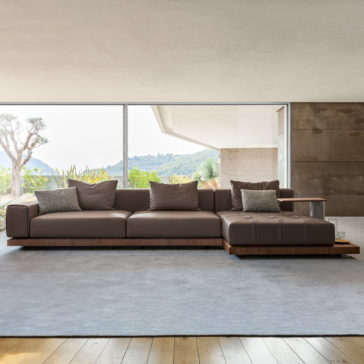 Contemporary High End Modular Corner Sofa A modern living room featuring the Contemporary High End Modular Corner Sofa in rich brown leather, adorned with multiple cushions, set on a light gray rug. Behind the sofa, a large window reveals a scenic view of lush green hills and a clear sky. The room boasts a combination of light and dark neutral tones.