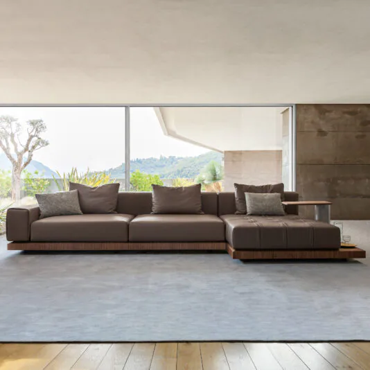 A modern living room featuring the Contemporary High End Modular Corner Sofa in rich brown leather, adorned with multiple cushions, set on a light gray rug. Behind the sofa, a large window reveals a scenic view of lush green hills and a clear sky. The room boasts a combination of light and dark neutral tones.