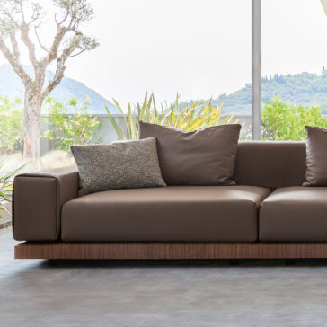 Contemporary High End Modular Corner Sofa A Contemporary High End Modular Corner Sofa, upholstered in elegant brown leather with a sleek wooden base, graces a bright room filled with natural light from large windows. The chic design showcases two spacious back cushions and a smaller decorative pillow for added style. Beyond the windows, lush green foliage and rolling hills stretch under a brilliant sky.