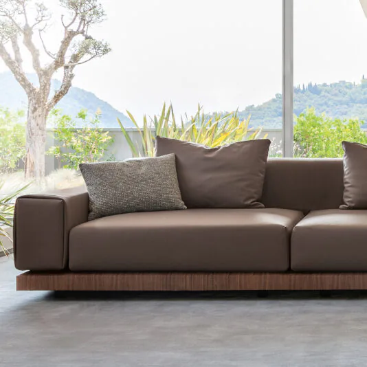 A Contemporary High End Modular Corner Sofa, upholstered in elegant brown leather with a sleek wooden base, graces a bright room filled with natural light from large windows. The chic design showcases two spacious back cushions and a smaller decorative pillow for added style. Beyond the windows, lush green foliage and rolling hills stretch under a brilliant sky.