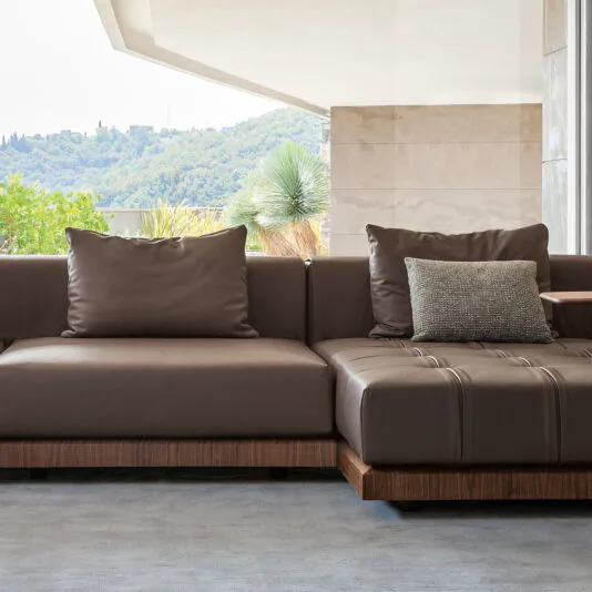 The modern, spacious living room features a Contemporary High End Modular Corner Sofa, adorned with cushions. This L-shaped brown leather sofa enhances the room, which includes an open wall offering a view of green hills and trees, providing a relaxing and airy atmosphere. The light gray flooring complements the elegant decor.