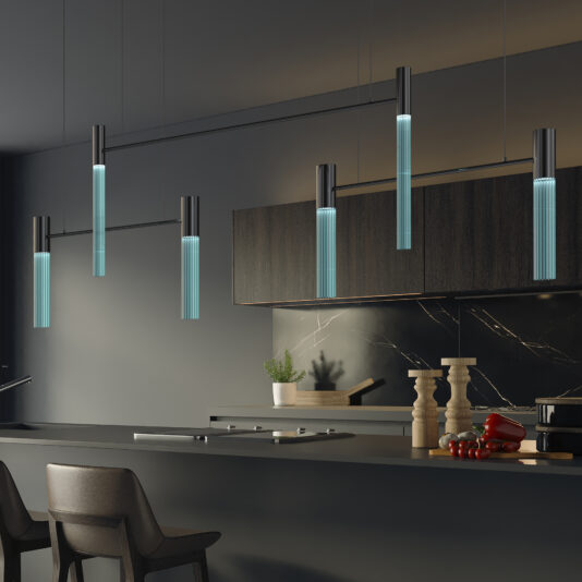 A contemporary kitchen showcases a sleek black island paired with high chairs. The star of the design is the Contemporary Modular Glass Tube Chandelier hanging above, casting a beautiful translucent glow. Complementing this elegant setup are dark cabinetry, a marble backsplash, and decor elements including a few vegetables, a potted plant, and spice mills on the counter.