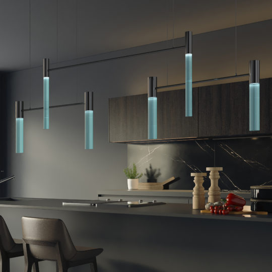 A contemporary kitchen showcases a sleek black island paired with high chairs. The star of the design is the Contemporary Modular Glass Tube Chandelier hanging above, casting a beautiful translucent glow. Complementing this elegant setup are dark cabinetry, a marble backsplash, and decor elements including a few vegetables, a potted plant, and spice mills on the counter.