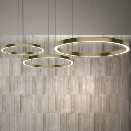 Three large, circular gold ring light fixtures, the Contemporary Round Downlight Chandelier, hang from the ceiling against a backdrop of light wooden wall panels. These chandeliers are suspended at varying heights, creating a modern and minimalistic look.