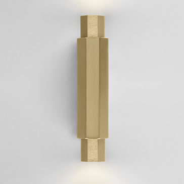 Geometric Satin Gold Tube Style Wall Light The Geometric Satin Gold Tube Style Wall Light emits soft light from both ends. This modern fixture boasts a sleek, hexagonal shape and a brushed metal finish, enhancing its minimalist aesthetic when mounted on a plain white wall.