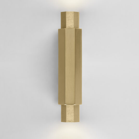 Geometric Satin Gold Tube Style Wall Light The Geometric Satin Gold Tube Style Wall Light emits soft light from both ends. This modern fixture boasts a sleek, hexagonal shape and a brushed metal finish, enhancing its minimalist aesthetic when mounted on a plain white wall.