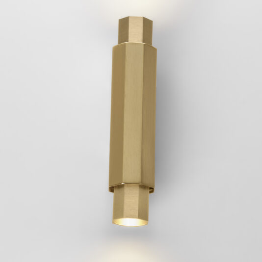 A stylish Geometric Satin Gold Tube Style Wall Light with a sleek, cylindrical design is mounted against a plain, light gray background. The fixture emits a soft white light from both ends, creating an elegant and minimalist aesthetic.