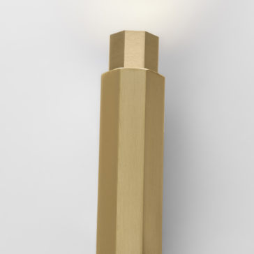 Geometric Satin Gold Tube Style Wall Light A close-up image of a modern, geometric, satin gold-colored hexagonal wall light with smooth, flat sides. The wall light is designed in a minimalist style and is shown against a plain, light grey background. The upper part of the fixture is illuminated.