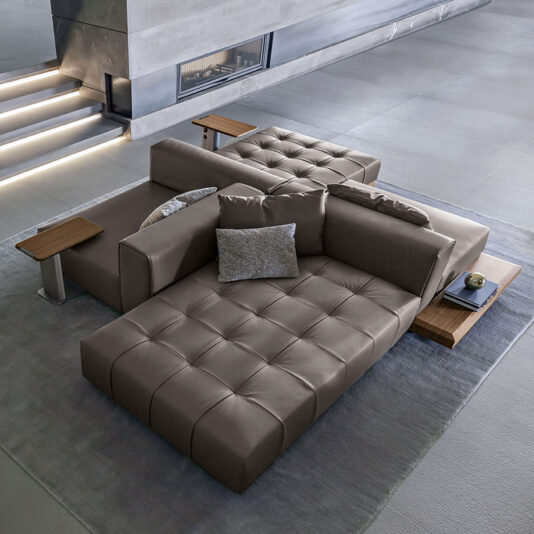 High End Contemporary Modular Sofa A sophisticated, low-profile modular sofa from the High End Contemporary Modular Sofa collection features tufted leather cushions in a minimalist living room. This premium sectional is complemented by sleek wooden side tables and stylish decorative cushions. The space showcases large grey tiles and a staircase with ambient lighting in the background.