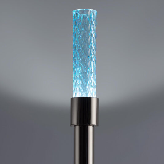 The Modern Faceted Crystal Floor Lamp, designed with a slim cylindrical shape and sleek metallic base, features a textured glass tube. The intricately patterned faceted glass is softly illuminated with blue light, enhancing its sophisticated and contemporary appearance against a neutral gray background.