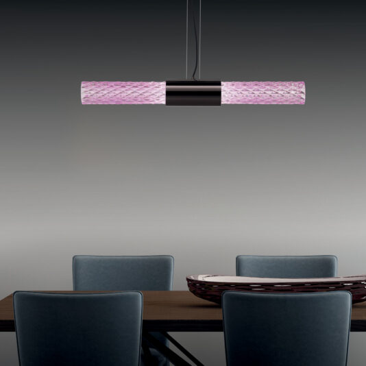 A Modern Faceted Crystal Horizontal Pendant Light with a pink patterned shade illuminates a wooden dining table, which is paired with four dark blue upholstered chairs. The table showcases a decorative bowl with a glossy finish, all set against a gradient gray background wall.