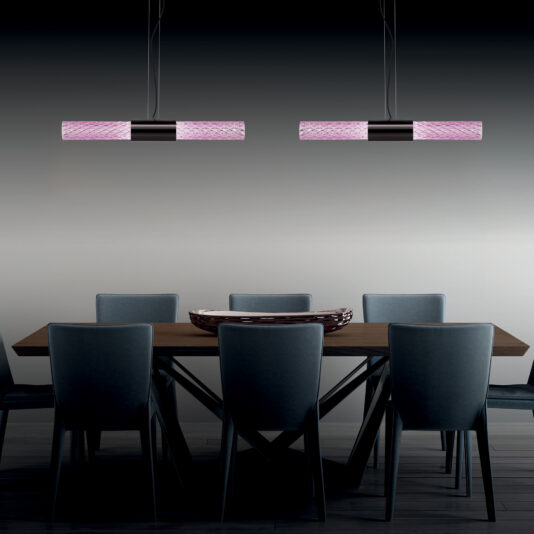 Modern Faceted Crystal Horizontal Pendant Light A modern dining room features a wooden table surrounded by six gray upholstered chairs. Above the table, two Modern Faceted Crystal Horizontal Pendant Lights with pink and black accents hang from the ceiling, creating a minimalist and stylish ambiance.