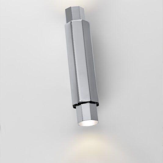 The Modern Geometric Tube Style Wall Light, sporting a sleek, metallic finish, is mounted on a white wall. This angularly designed fixture emits light from both ends—one directed upwards and the other downwards—crafting a sophisticated ambiance.
