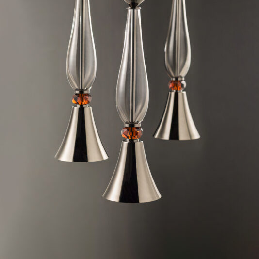 A close-up view of the Modern Pendant Light With Cut Crystal Detail showcases three sleek, metallic pendants with elongated, tapered tops and bell-shaped covers. Each pendant includes a small orange accent near the top. The background is a smooth, dark gradient.
