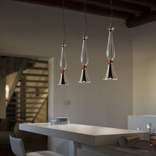 A modern kitchen showcases three Modern Pendant Lights With Cut Crystal Detail elegantly hanging over a white island counter. The pendant lights feature teardrop-shaped glass shades with metal accents. In the background, a staircase leads to a cozy living area, and the overall scene is softly lit, creating a warm atmosphere.