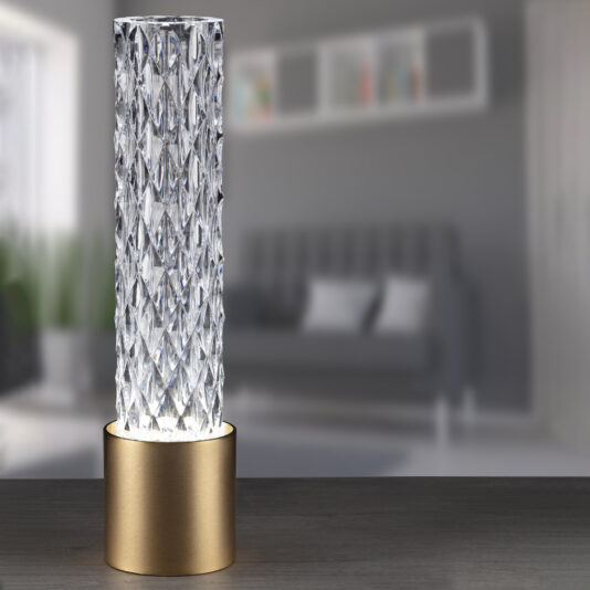 The Modern Vertical Faceted Crystal Table Lamp, with its luxurious cylindrical design and intricate diamond patterns, stands elegantly on a wooden surface. Its base features a stunning gold-colored material. The background showcases a blurred living room scene complete with a gray sofa and wall-mounted shelves.