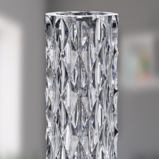 Modern Vertical Faceted Crystal Table Lamp A close-up of a tall, cylindrical faceted vase with intricate interlocking diamond patterns. The background is blurred, highlighting the vase's detailed and reflective surface, reminiscent of a Modern Vertical Faceted Crystal Table Lamp.