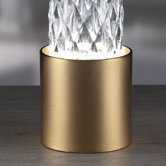 Modern Vertical Faceted Crystal Table Lamp A close-up shot of the Modern Vertical Faceted Crystal Table Lamp exhibits its cylindrical base with a smooth, metallic gold finish. The top part, crafted from transparent faceted crystal, radiates a bright light that highlights the intricate pattern of the glass.