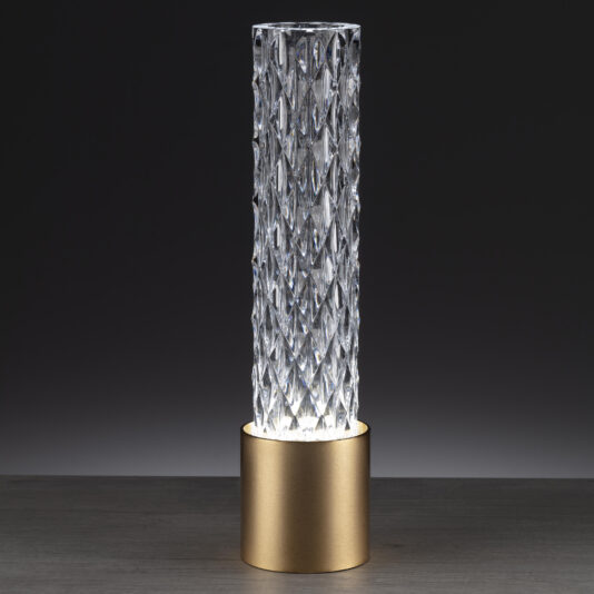 Modern Vertical Faceted Crystal Table Lamp The image showcases the Modern Vertical Faceted Crystal Table Lamp, featuring a textured cylindrical glass design that emits light. The glass sits in a matte gold metal base, set against a dark backdrop that highlights the clear, faceted crystal patterns of the lamp.