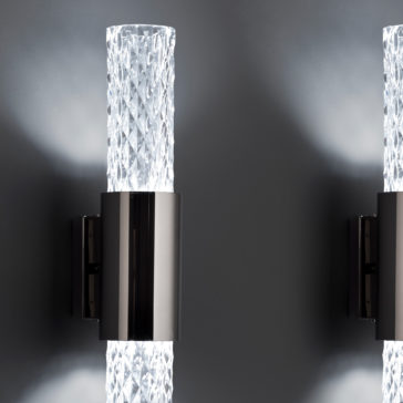 Modern Vertical Faceted Crystal Wall Light Close-up of two Modern Vertical Faceted Crystal Wall Lights with sleek metallic bases. The sconces are illuminated, casting a soft glow against a dark gray wall, highlighting their intricate crystal-textured glass cylinders and contemporary design.