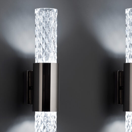 Modern Vertical Faceted Crystal Wall Light Close-up of two Modern Vertical Faceted Crystal Wall Lights with sleek metallic bases. The sconces are illuminated, casting a soft glow against a dark gray wall, highlighting their intricate crystal-textured glass cylinders and contemporary design.