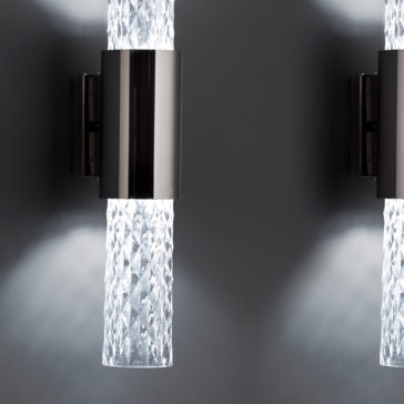 Modern Vertical Faceted Crystal Wall Light Close-up of two Modern Vertical Faceted Crystal Wall Lights featuring cylindrical, diamond-patterned glass shades extending from brushed metal mounts. The lights are on, casting a soft glow against a smooth grey wall.