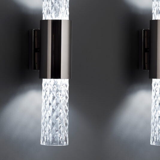 Modern Vertical Faceted Crystal Wall Light Close-up of two Modern Vertical Faceted Crystal Wall Lights featuring cylindrical, diamond-patterned glass shades extending from brushed metal mounts. The lights are on, casting a soft glow against a smooth grey wall.