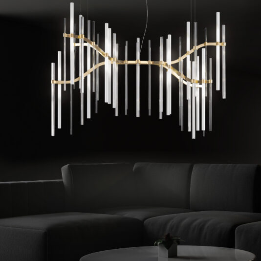 A Vertical Murano Glass Tube Modern Chandelier with multiple vertical glass tubes illuminates against a dark background, hanging over a grey sectional sofa. The lighting fixture's undulating gold frame adds a decorative and sophisticated ambiance to the setting.