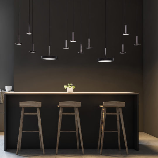 A modern kitchen with a dark wall features three wooden bar stools lined up at a central island. Above the island, **Contemporary Round Pendant Lights** of varying heights hang from the ceiling. A small potted plant sits on the island, adding a touch of greenery.