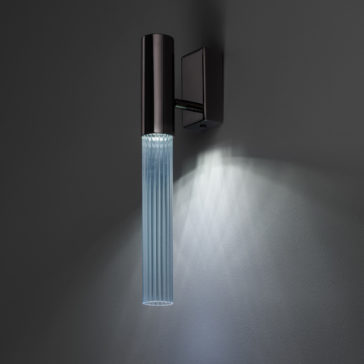Against a dark grey background, the Contemporary Vertical Glass Tube Wall Light—a modern cylindrical black sconce with vertical ridges—emits a soft, diffused light. The pattern of rays it creates on the wall below enhances its sleek and contemporary design.
