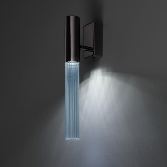 Against a dark grey background, the Contemporary Vertical Glass Tube Wall Light—a modern cylindrical black sconce with vertical ridges—emits a soft, diffused light. The pattern of rays it creates on the wall below enhances its sleek and contemporary design.