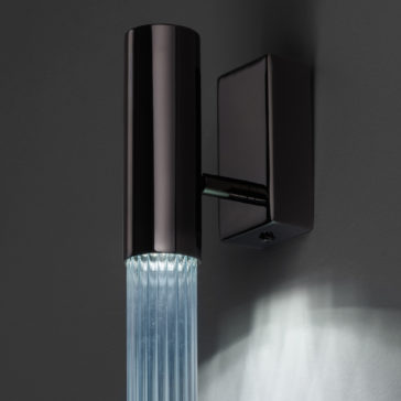 The Contemporary Vertical Glass Tube Wall Light boasts a sleek, modern design with a cylindrical black metal casing and a clear ribbed glass tube beneath it, beautifully casting soft white light onto the gray wall behind it.