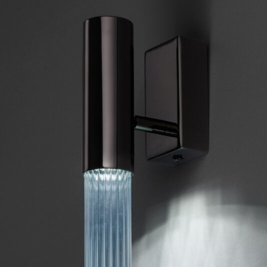 Contemporary Vertical Glass Tube Wall Light The Contemporary Vertical Glass Tube Wall Light boasts a sleek, modern design with a cylindrical black metal casing and a clear ribbed glass tube beneath it, beautifully casting soft white light onto the gray wall behind it.