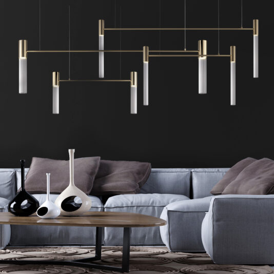 A modern living room with a dark wall and contemporary lighting fixtures, including a Large Contemporary Modular Glass Tube Chandelier, hanging from the ceiling. There is a grey sectional sofa with neutral pillows and a wooden coffee table adorned with various artistic vases in black and white. The floor has a patterned rug.