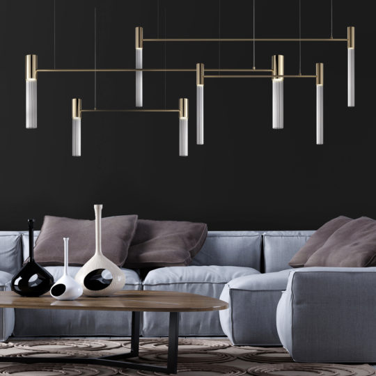 A modern living room with a dark wall and contemporary lighting fixtures, including a Large Contemporary Modular Glass Tube Chandelier, hanging from the ceiling. There is a grey sectional sofa with neutral pillows and a wooden coffee table adorned with various artistic vases in black and white. The floor has a patterned rug.