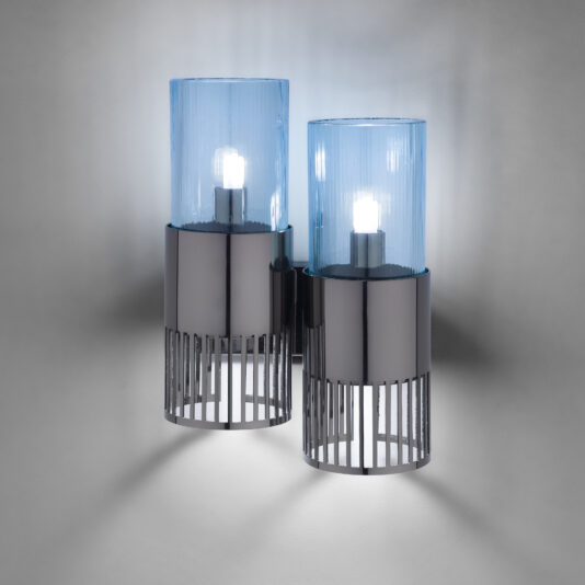 Modern Double Wall Light With Glass Shades The Modern Double Wall Light With Glass Shades features two cylindrical blue glass covers, each housing a lit bulb. These shades are mounted on a sleek metallic base with vertical slits, which casts a soft ambient light against the background. The design is contemporary and minimalistic.