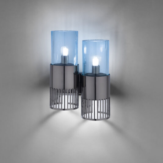 Introducing the Modern Double Wall Light With Glass Shades: featuring two sleek cylindrical lights, each with a metallic base adorned with vertical slits and topped with a blue glass shade. These lights emit a soft glow that casts intriguing shadows on the wall, creating a contemporary and stylish ambiance.