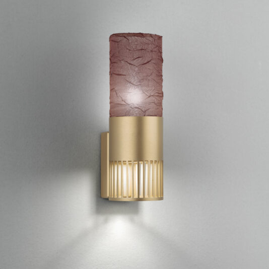 The Modern Single Wall Light With Shade features a sleek, textured, cylindrical shade in a rich reddish-brown hue, mounted on an elegant gold-toned base with vertical slits. This fixture casts a warm light against a plain grey wall, seamlessly blending contemporary design with subtle elegance.