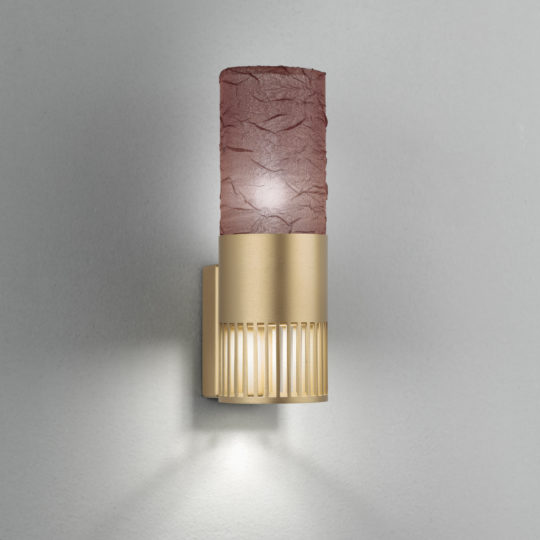 The Modern Single Wall Light With Shade features a sleek, textured, cylindrical shade in a rich reddish-brown hue, mounted on an elegant gold-toned base with vertical slits. This fixture casts a warm light against a plain grey wall, seamlessly blending contemporary design with subtle elegance.