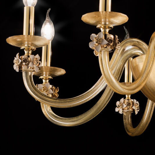 A close-up of the 8 Arm Hand Blown Glass Chandelier With Flowers shows its ornate design featuring four candle-like light bulbs. The chandelier boasts curved, gilded arms adorned with small, intricate floral decorations. Set against a black background, the chandelier's details and warm glow are beautifully highlighted.