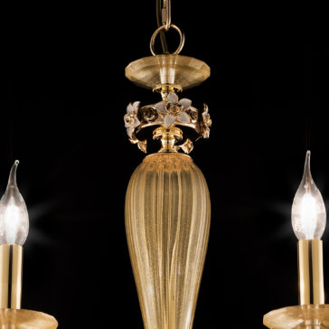 8 Arm Hand Blown Glass Chandelier With Flowers A close-up view of the 8 Arm Hand Blown Glass Chandelier With Flowers highlights its ornate gold finish and a central vertical piece adorned with intricate floral designs. Two light bulbs, featuring flame-shaped covers, cast a warm glow that accentuates the exquisite golden details against a dark backdrop.