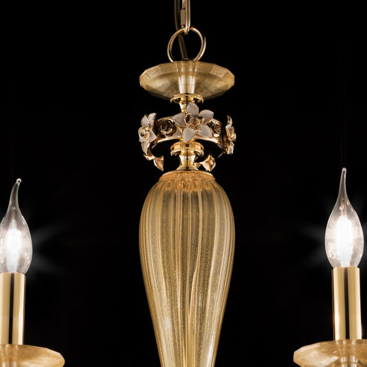 A close-up view of the 8 Arm Hand Blown Glass Chandelier With Flowers highlights its ornate gold finish and a central vertical piece adorned with intricate floral designs. Two light bulbs, featuring flame-shaped covers, cast a warm glow that accentuates the exquisite golden details against a dark backdrop.