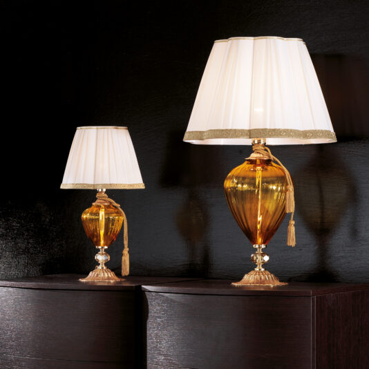 Two Amber Glass Table Lamps with cut crystal detail and white pleated lampshades, featuring gold trim and adorned with gold tassels, are displayed on a dark wooden surface against a black background. The smaller lamp is positioned slightly behind the larger one.