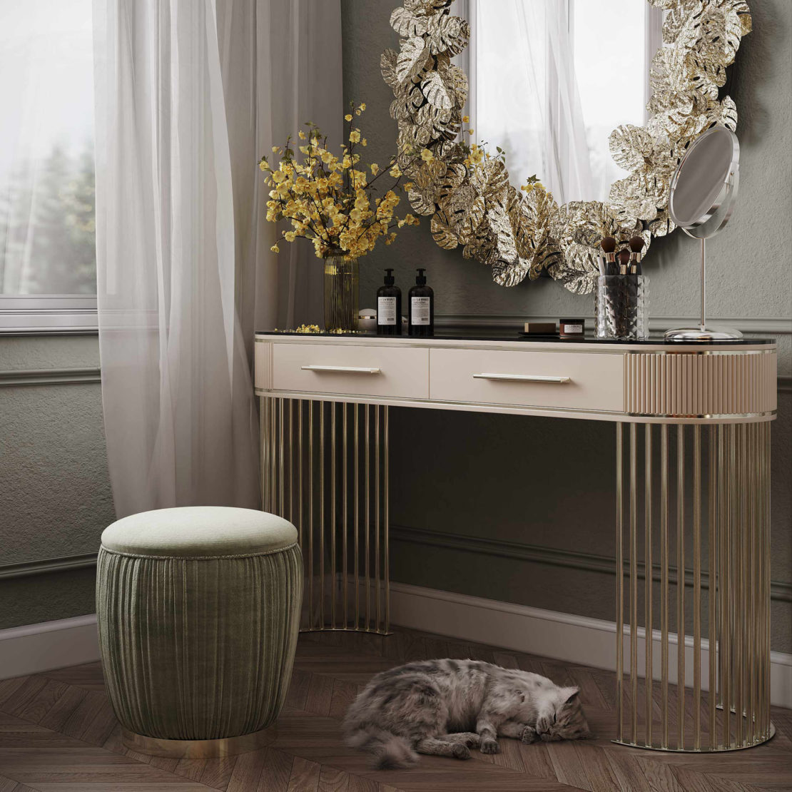 A chic vanity setup features an Art Deco Style Ribbed Dressing Table with gold accents and two drawers, a round cushioned stool, a large ornate mirror with a leaf motif frame, and a tabletop adorned with beauty products and a vase of yellow flowers. A cat sleeps on the herringbone-patterned floor.