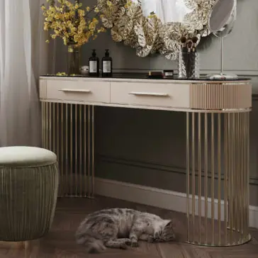 Art Deco Style Ribbed Dressing Table An Art Deco Style Ribbed Dressing Table with metallic legs and two drawers, decorated with yellow flowers, bottles, and a mirror. A round green ottoman is positioned beside the table, while a gray cat naps on the wooden floor beneath it.