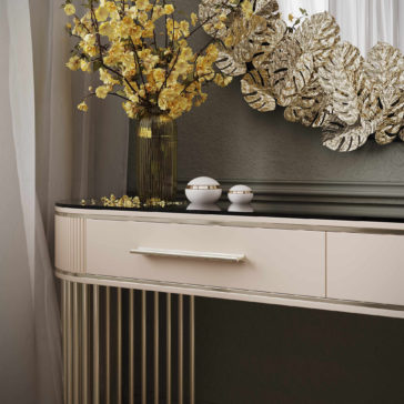 Art Deco Style Ribbed Dressing Table A stylish Art Deco Style Ribbed Dressing Table featuring a cream-colored finish with a reflective black top and vertical slatted legs is adorned with a tall green vase filled with yellow flowers, two small white spherical decorations, and a large decorative mirror accented with gold leaf.