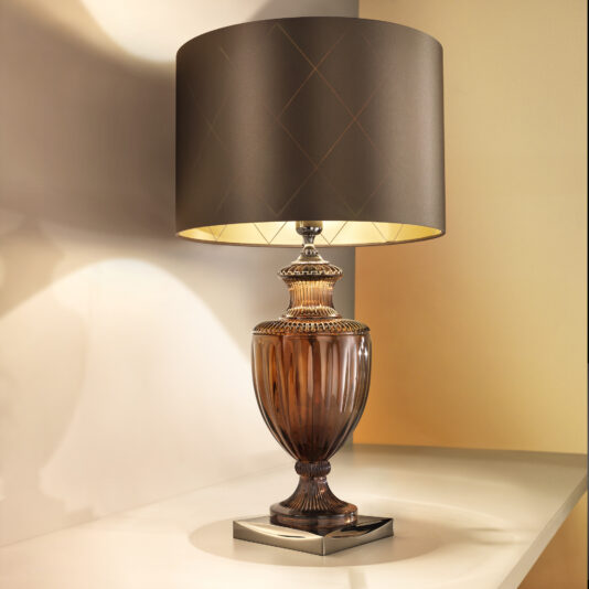 The Classic Style Brown Crystal Table Lamp, featuring an ornate amber-colored glass base and a large dark brown lampshade, sits on a reflective surface. It casts a soft, warm light and gentle shadows on the surrounding beige wall, exuding an elegant and timeless aesthetic.